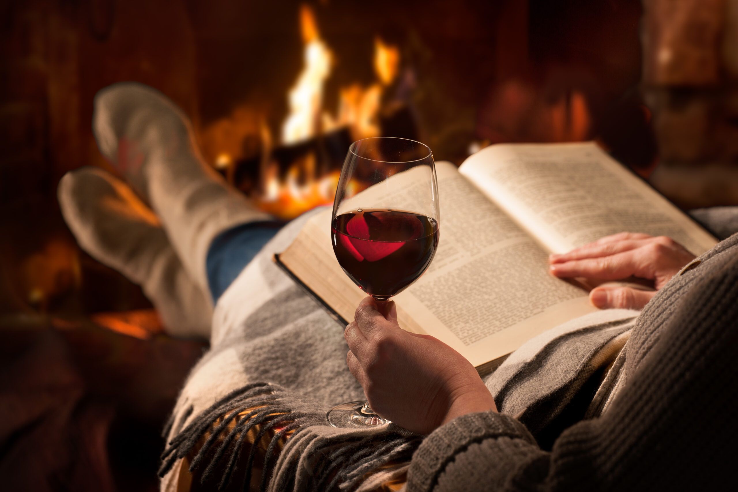 Una joven leyendo un libro con una copa de vino en la mano cerca de la chimenea